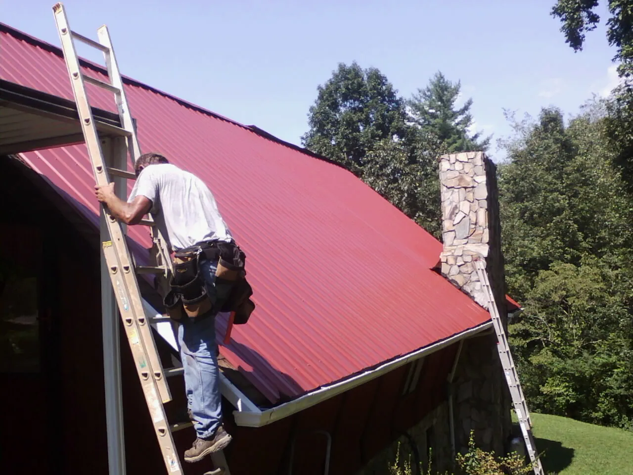 Roofer on a ladder beside a red metal roof and stone chimney — Soffit & Fascia Repair by Roofing Brothers in Silverlake
